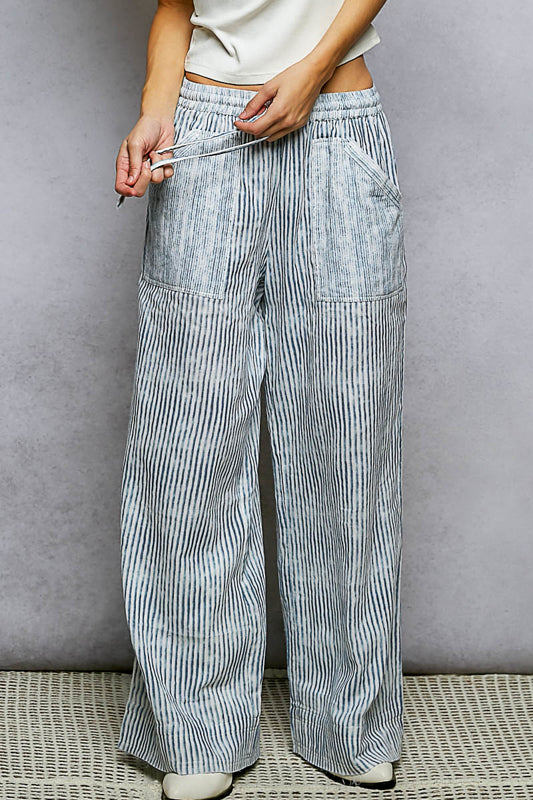 Front view of woman pulling the drawstring waist of light blue and white vertical stripe wide-leg pants with patch pockets, paired with a white crop top, on a gray background.