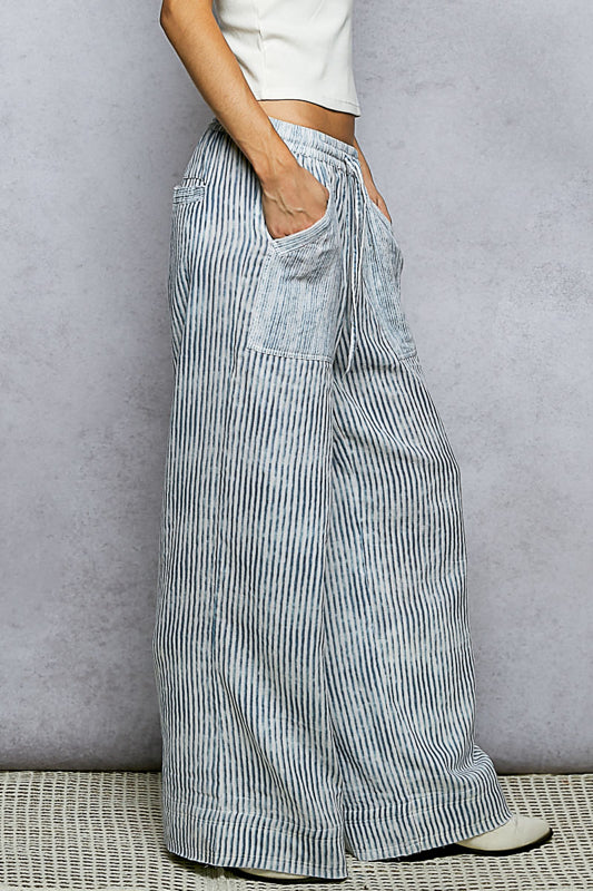 Side view of woman with hand in pocket wearing light blue and white vertical stripe wide-leg pants with patch pockets, paired with a white crop top, on a gray background.