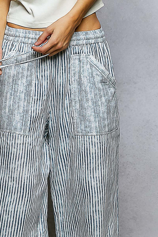 Close-up of the drawstring elastic waist and patch pocket detail on light blue and white vertical stripe wide-leg pants, on a gray background.