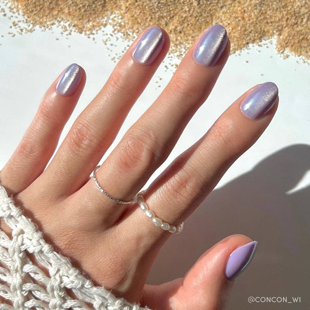 Close-up of hand wearing Glamnetic Mermaid's Tail Press-On Nails featuring short oval iridescent lavender chrome nails, styled with delicate gold and pearl stacked rings against a sandy beach background