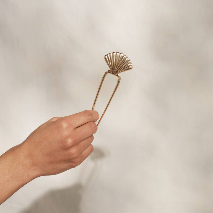 Hand holding a gold scallop shell two-prong hair pin against a neutral background.