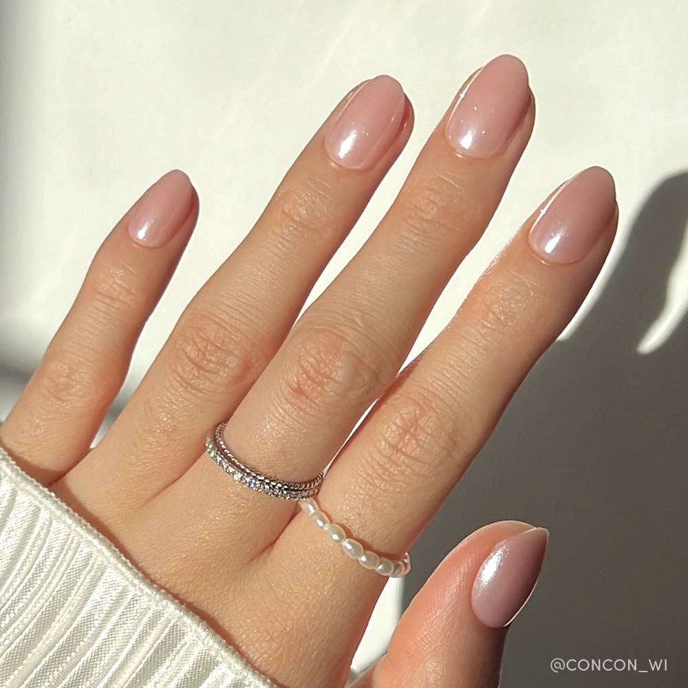 Close-up of blush pink short oval press-on nails with pearl shimmer finish worn on fair skin tone hand with silver and pearl stacked rings
