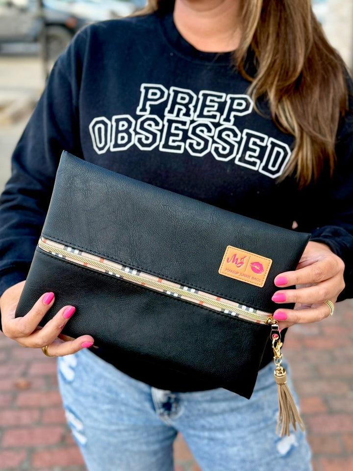 Woman in Prep Obsessed sweatshirt holding large black vegan leather Makeup Junkie zip pouch with plaid-trim gold zipper and tassel
