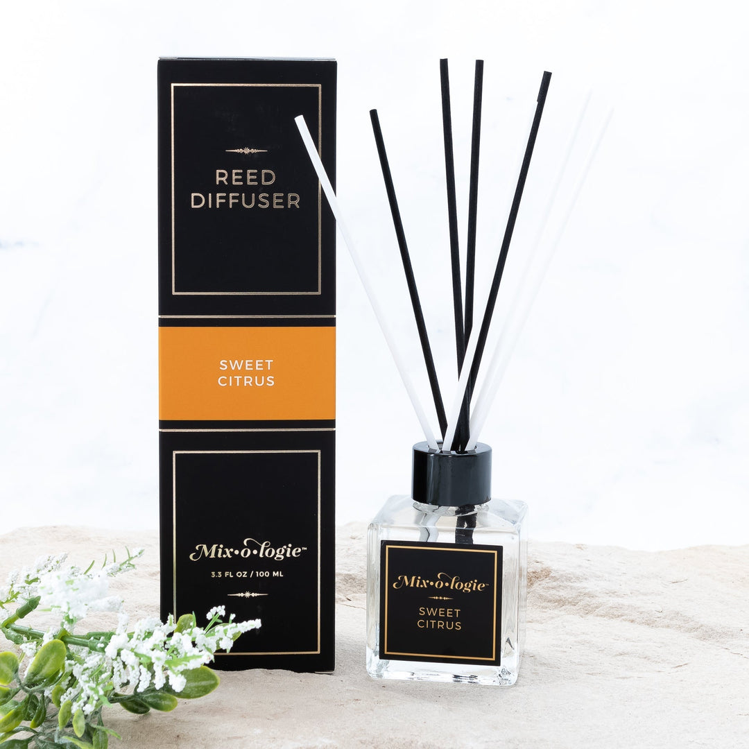 Mixologie Reed Diffuser in Sweet Citrus, 3.3 fl oz, clear square glass bottle with black cap and black label with orange accent band, with matching black and orange box, styled with white florals