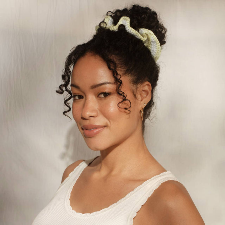Front view of a smiling woman with curly black hair in a high bun secured with the butter yellow and white crochet scrunchie, wearing a white tank top and gold hoop earrings.