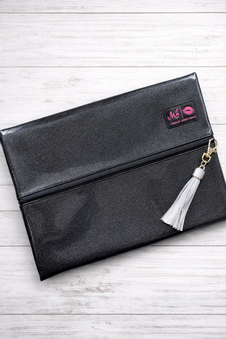 Makeup Junkie Bags grey charcoal stardust glitter foldover zip makeup bag closed with silver tassel zipper pull and branded patch, front view on white wood background