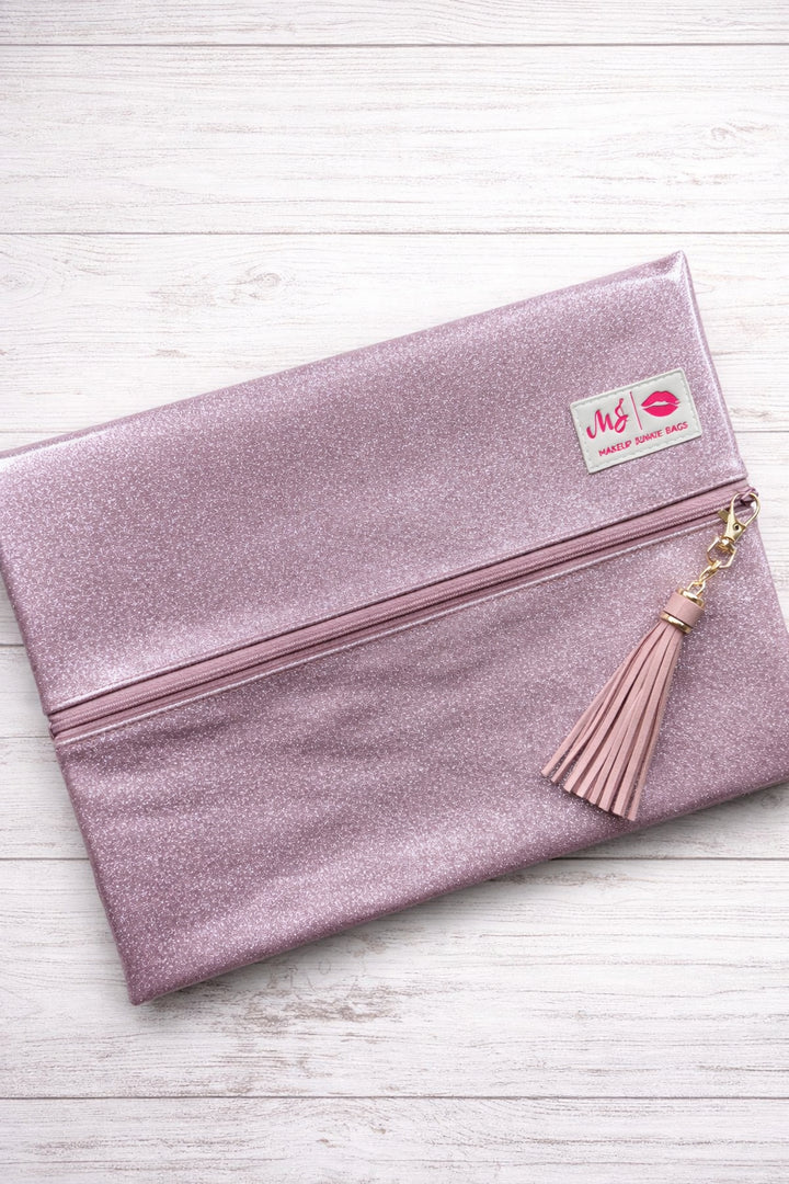 Makeup Junkie Bags blush pink glitter foldover zip makeup bag closed with blush tassel zipper pull and branded patch, front view on white wood background