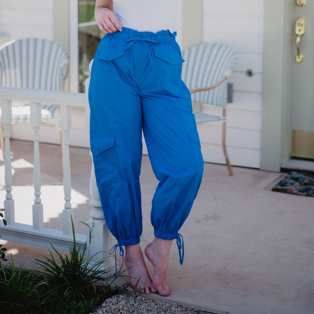 Woman on a porch stepping up on a stone, wearing a white top and cobalt blue paperbag-waist cargo jogger pants with tie ankles and side cargo pockets.