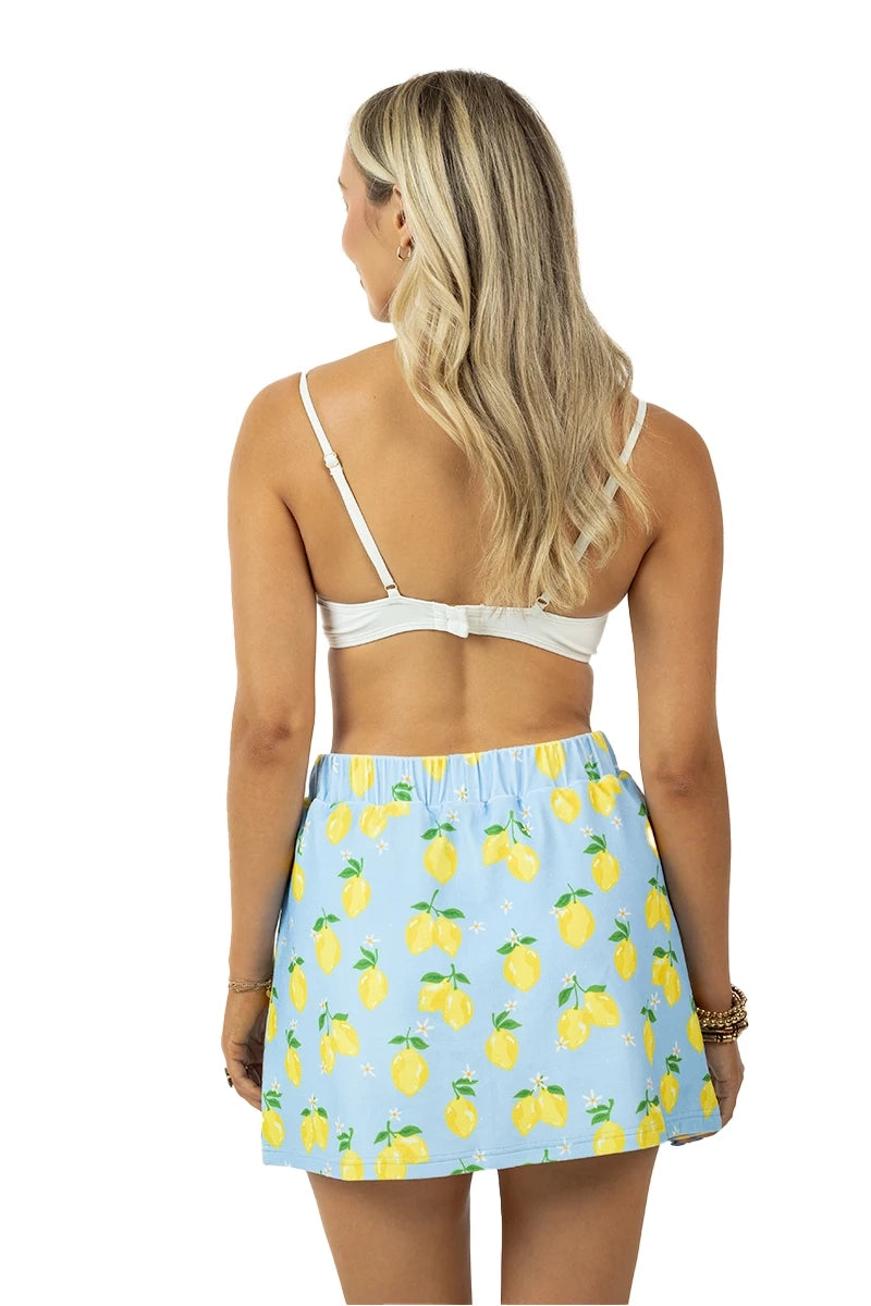 Back view of woman in a white bralette and light blue lemon print elastic-waist mini skirt with yellow lemons and green leaves, on a white background.