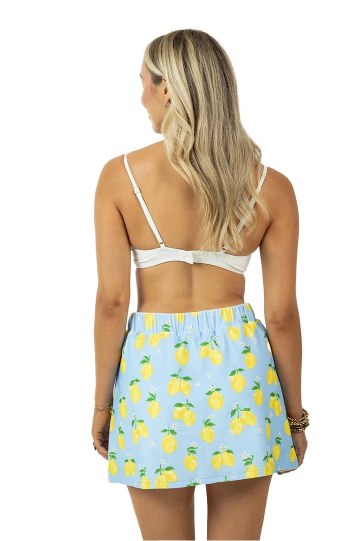 Back view of woman in a white bralette and light blue lemon print elastic-waist mini skirt with yellow lemons and green leaves, on a white background.