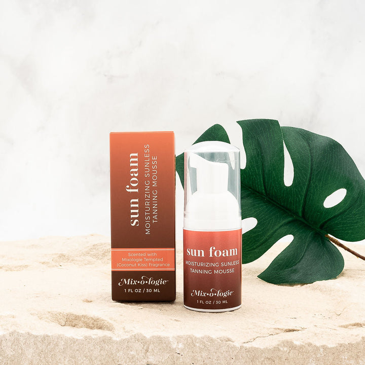 Mixologie Sun Foam Sunless Tanning Mousse 1 fl oz travel size bottle with matching brown and orange box styled on sandy surface with monstera leaf