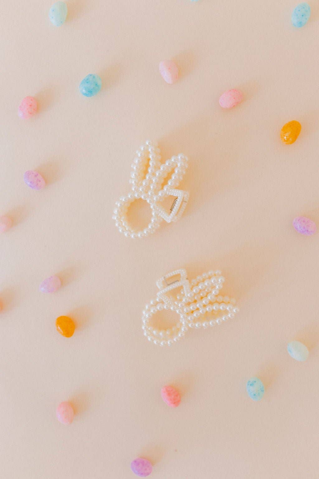 Two pearl beaded bunny ear claw clips flat lay on a blush pink surface scattered with pastel candy hearts in pink, blue, orange, and yellow.