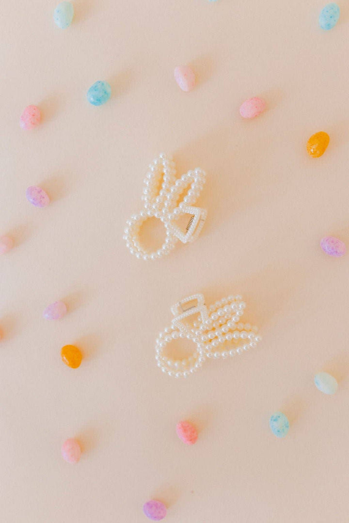 Two pearl beaded bunny ear claw clips flat lay on a blush pink surface scattered with pastel candy hearts in pink, blue, orange, and yellow.