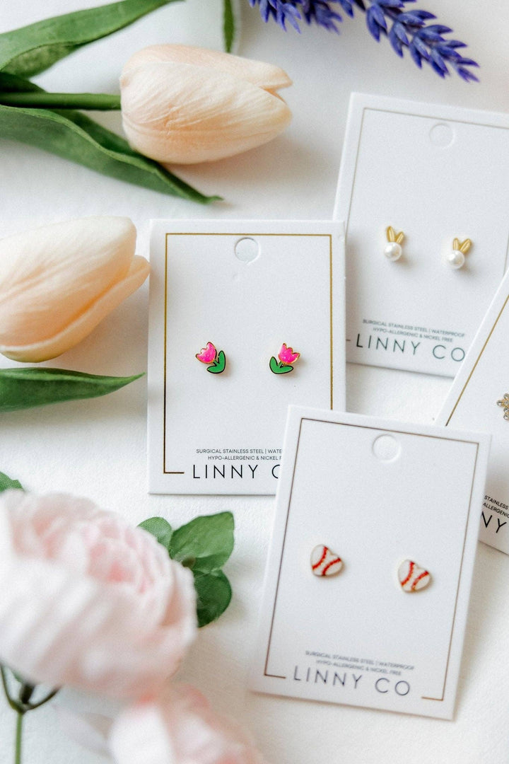 Three Linny Co stud earring cards flat lay surrounded by peach tulips, pink peonies, and purple lavender — pink tulip enamel studs in foreground, gold bunny ear pearl studs and red and white baseball enamel studs behind.