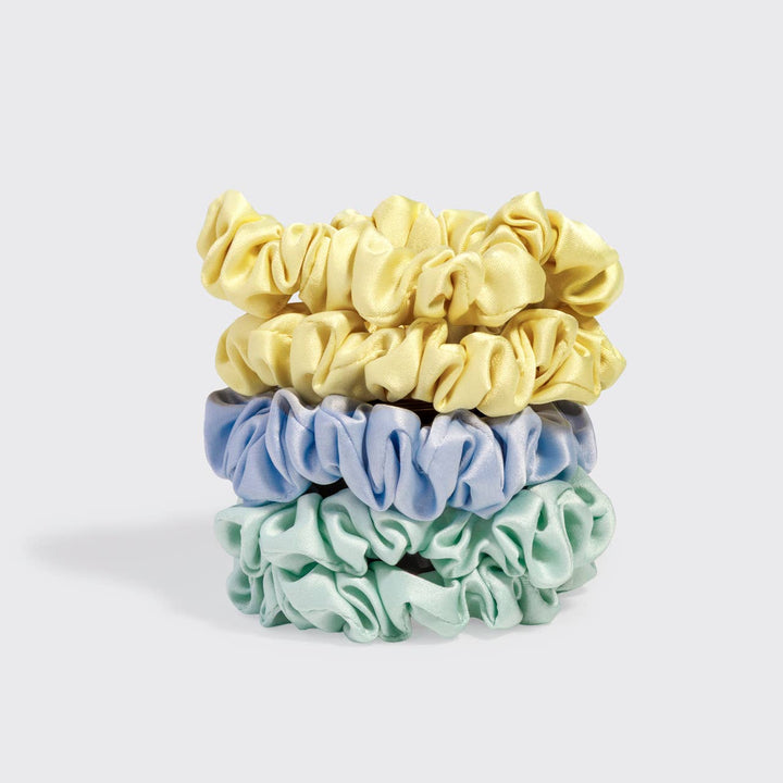 Stack of four ruched satin scrunchies in butter yellow (two), periwinkle blue, and mint green, on a white background.