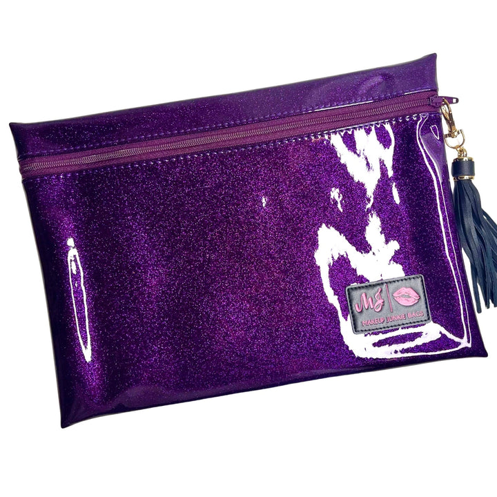 Makeup Junkie Bags deep purple glitter clear vinyl zip makeup bag with navy tassel zipper pull and branded patch, front view on white background