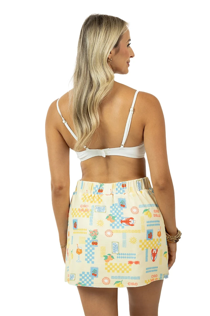 Back view of woman in a white bralette and cream Italian summer print elastic-waist mini skirt with lobster, lemon, and cocktail motifs, on a white background.