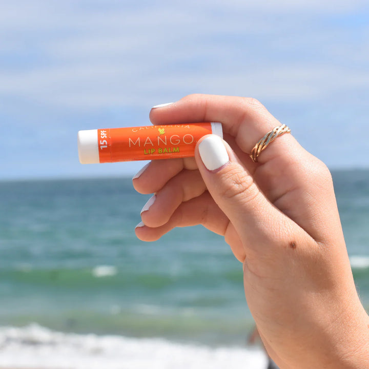 Hand holding California Mango SPF 15 Lip Balm stick at the beach with ocean and blue sky in background