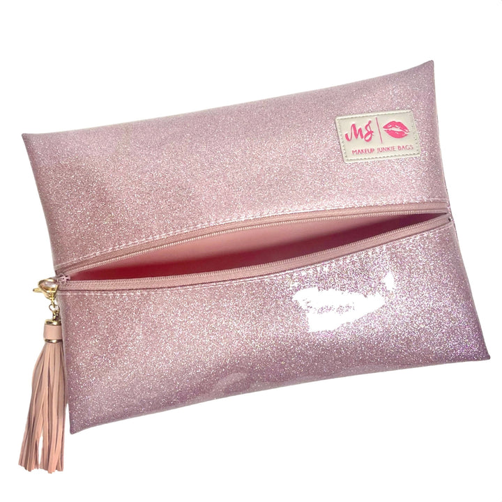 Makeup Junkie Bags blush pink glitter foldover zip makeup bag partially open showing hot pink interior lining with blush tassel zipper pull and branded patch, front view on white background