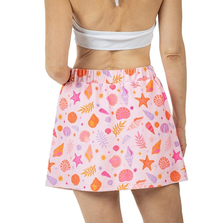 Back view of a pink seashell and starfish print elastic-waist mini skirt with orange, pink, and purple shell motifs, styled with a white bandeau top.