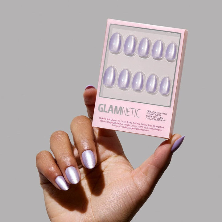 Hand holding Glamnetic Mermaid's Tail Press-On Nails pink packaging showing two rows of short oval iridescent lavender chrome nails against a gray background