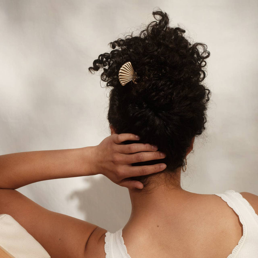 Back view of a woman with curly black hair in an updo, holding it in place with a gold scallop shell hair pin.
