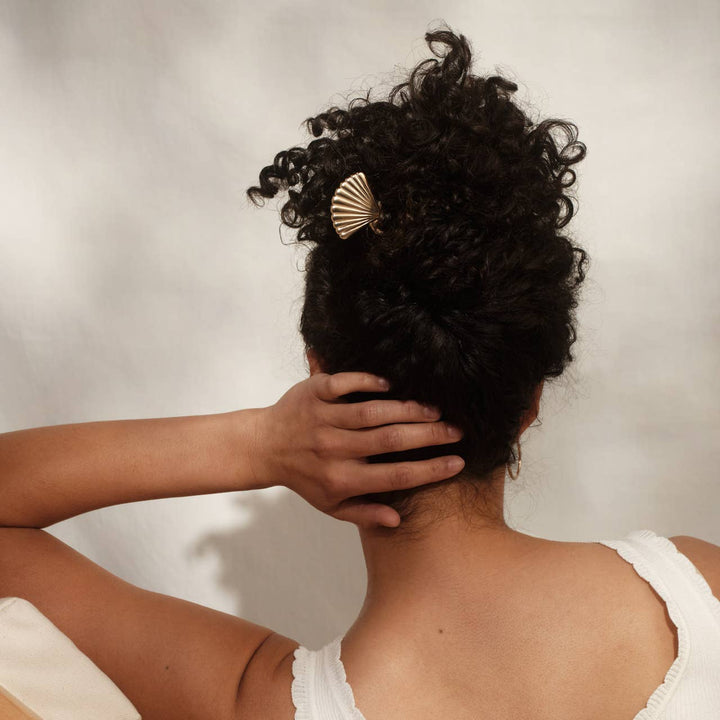 Back view of a woman with curly black hair in an updo, holding it in place with a gold scallop shell hair pin.
