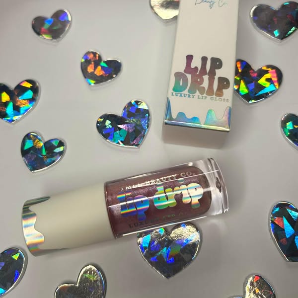 TMLL Beauty Co. Lip Drip Luxury Lip Gloss in a deep mauve shade with holographic packaging and gift box, surrounded by holographic heart confetti