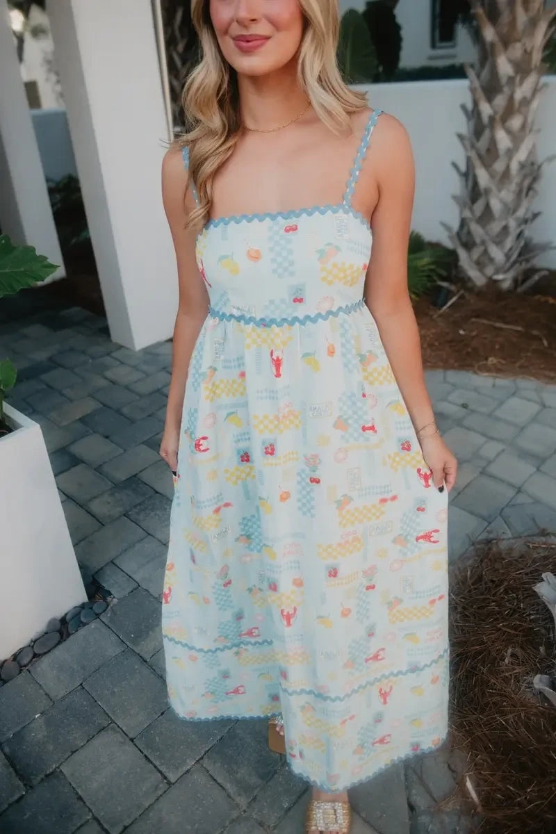 Woman smiling in a light blue lobster and summer print maxi dress with blue ric-rac trim, standing on a cobblestone patio with palm trees in the background.