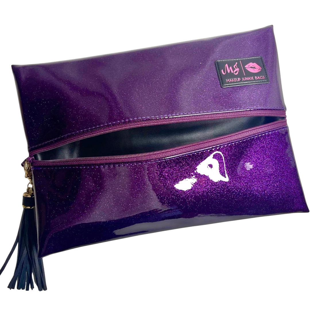 Makeup Junkie Bags deep purple glitter foldover zip makeup bag partially open with navy tassel zipper pull and branded patch, front view on white background