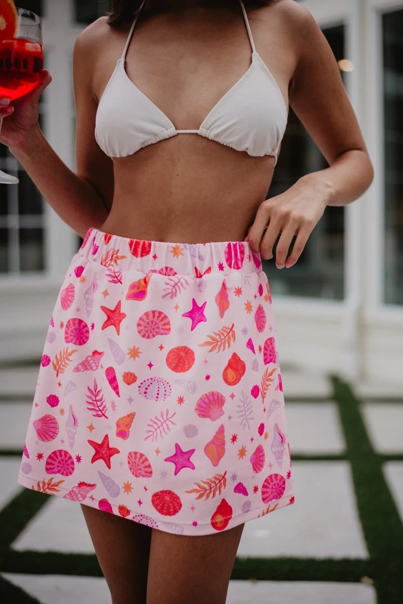 Close-up front view of a pink seashell and starfish print elastic-waist mini skirt with pink and red shell motifs, styled with a white bikini top outdoors.