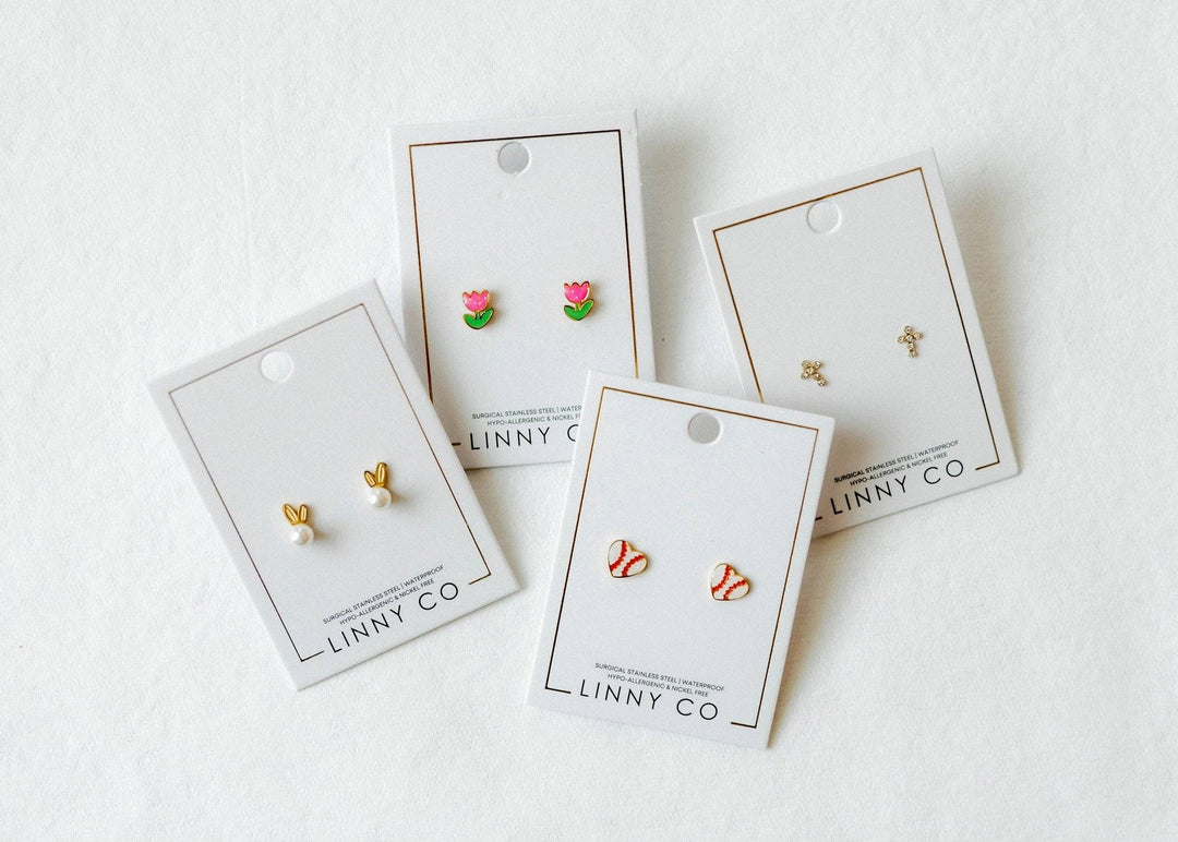 Four Linny Co stud earring cards flat lay on white background — pink tulip enamel studs, gold bunny ear pearl studs, red and white baseball enamel studs, and gold cross studs.