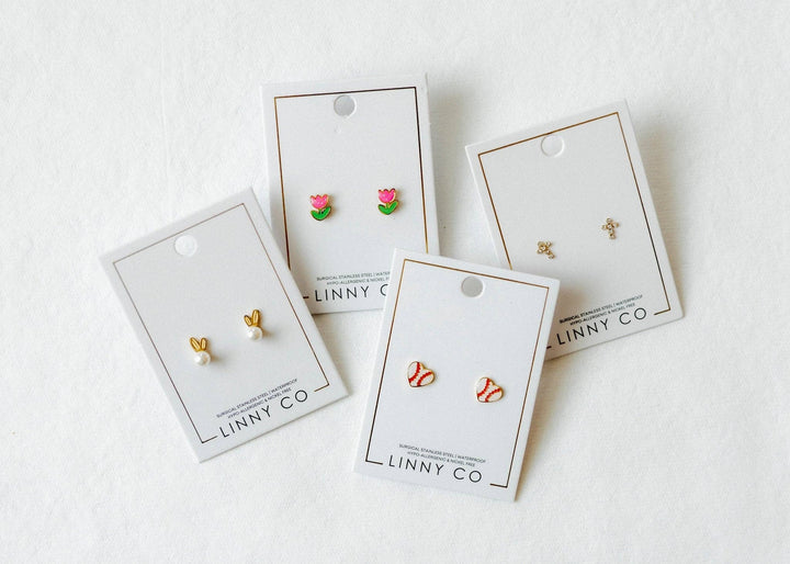 Four Linny Co stud earring cards flat lay on white background — pink tulip enamel studs, gold bunny ear pearl studs, red and white baseball enamel studs, and gold cross studs.