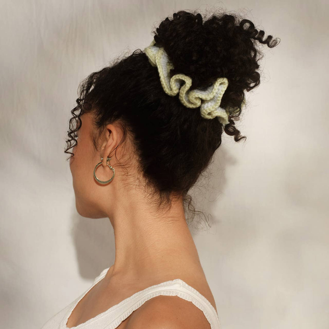 Side profile of a woman with curly black hair in a high bun secured with the butter yellow and white crochet scrunchie, wearing a white tank top and gold hoop earring.