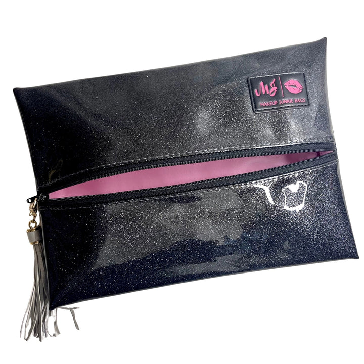 Makeup Junkie Bags grey charcoal stardust glitter foldover zip makeup bag partially open showing blush pink interior lining with silver tassel zipper pull and branded patch, front view on white background