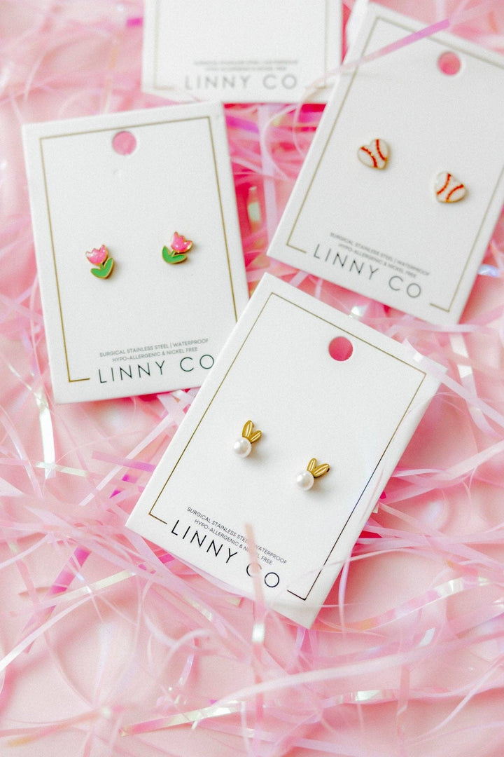 Three Linny Co stud earring cards flat lay on pink ribbon confetti background — pink tulip enamel studs, red and white baseball enamel studs, and gold bunny ear pearl studs.