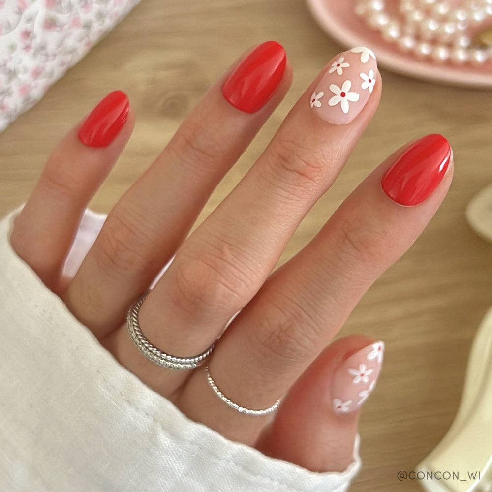 Close-up of hand wearing Glamnetic Red Blossom Press-On Nails featuring red short round nails with a nude accent nail decorated with white daisy flowers and a silver twisted ring