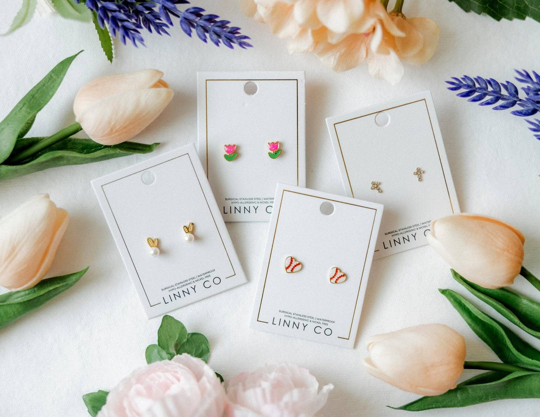 Four Linny Co stud earring cards flat lay surrounded by peach tulips, pink peonies, and purple lavender — gold bunny ear pearl studs, pink tulip enamel studs, red and white baseball enamel studs, and gold cross studs.
