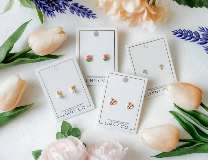 Four Linny Co stud earring cards flat lay surrounded by peach tulips, pink peonies, and purple lavender — gold bunny ear pearl studs, pink tulip enamel studs, red and white baseball enamel studs, and gold cross studs.