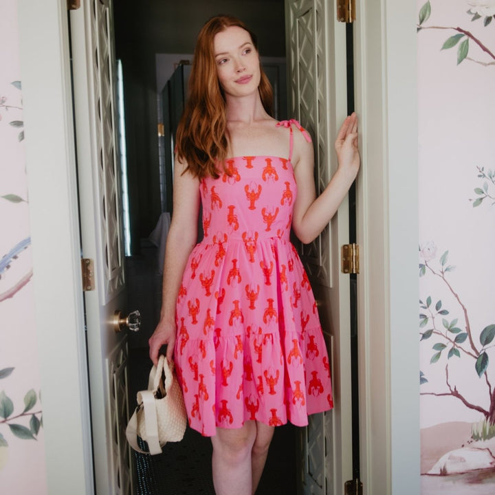 Woman with red hair standing in a doorway with floral wallpaper, wearing a pink orange lobster print tiered mini dress with tie straps, holding a white structured bag.