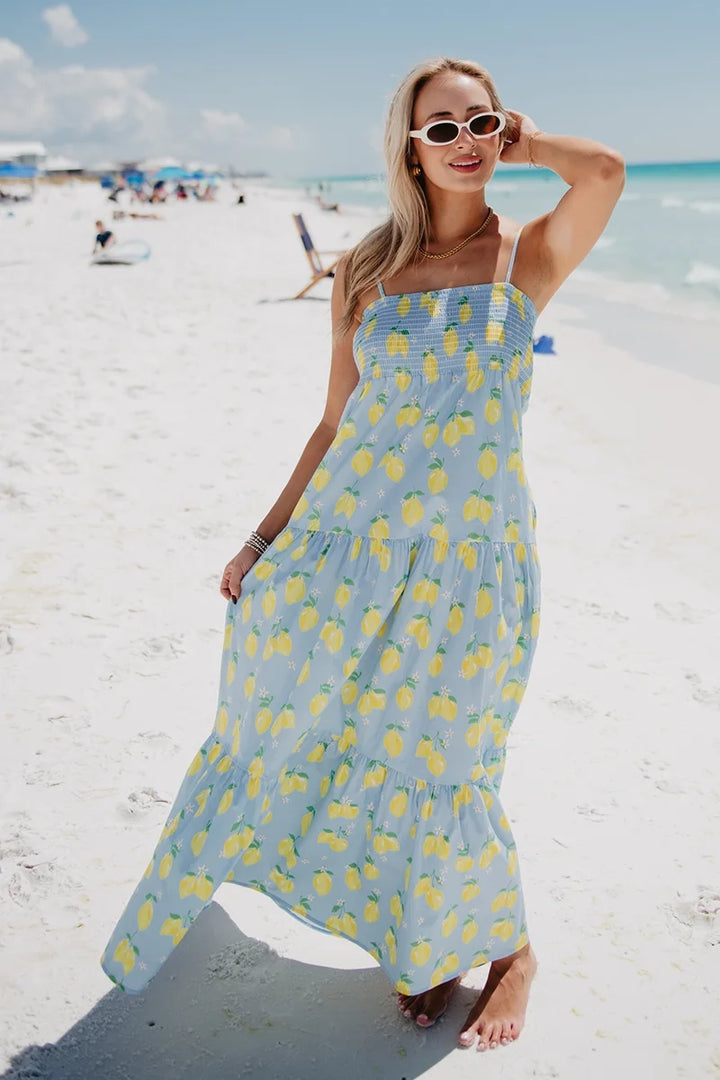 Woman in white oval sunglasses wearing a light blue lemon print smocked tiered maxi dress with hand raised to hair, standing on a white sand beach with turquoise water in the background.
