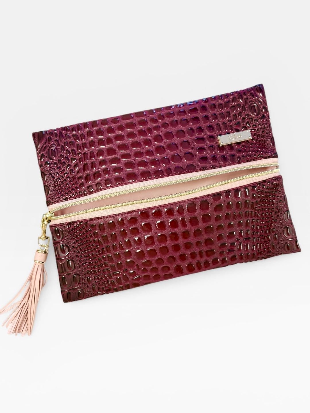 Burgundy crocodile embossed faux leather fold-over clutch with gold zipper and blush pink tassel pull, on a white background.