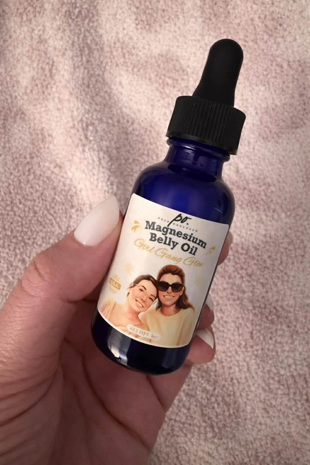 Hand holding a blue glass dropper bottle of Magnesium Belly Oil against a textured blush pink fabric background.