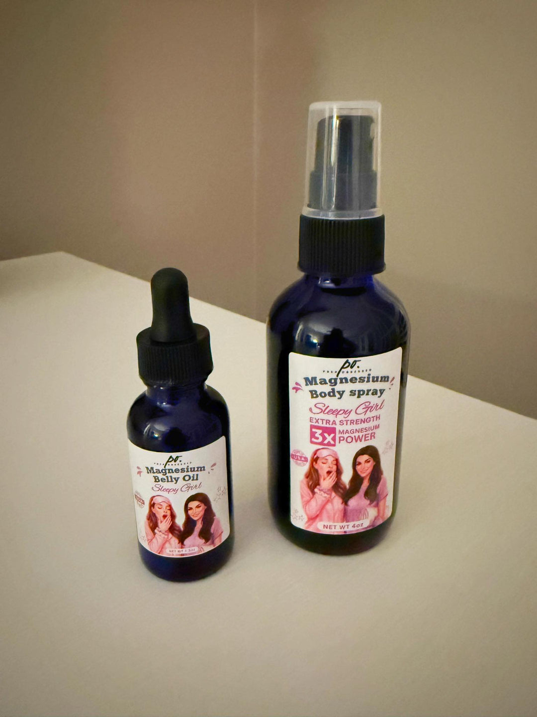 Magnesium Belly Oil dropper bottle and Magnesium Body Spray Extra Strength 3X in blue glass bottles displayed on a white surface against a neutral wall.