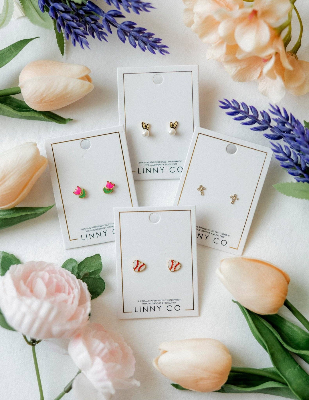Four Linny Co stud earring cards flat lay surrounded by peach tulips, pink peonies, and purple lavender — gold bunny ear pearl studs, pink tulip enamel studs, red and white baseball enamel studs, and gold cross studs.