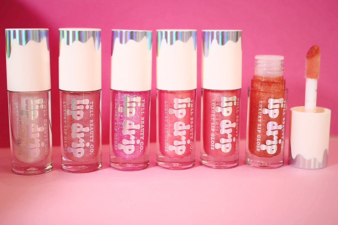 Six Lip Drip Luxury Lip Gloss tubes in sheer to bold pink and copper shades displayed on a pink background, one open showing applicator wand