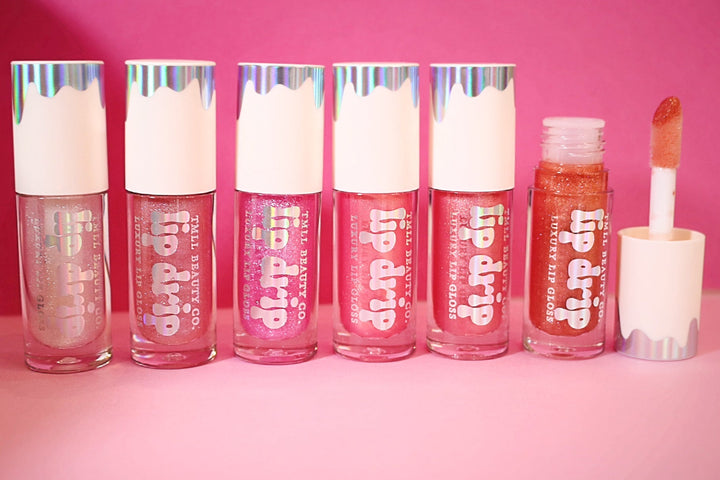 Six Lip Drip Luxury Lip Gloss tubes in sheer to bold pink and copper shades displayed on a pink background, one open showing applicator wand