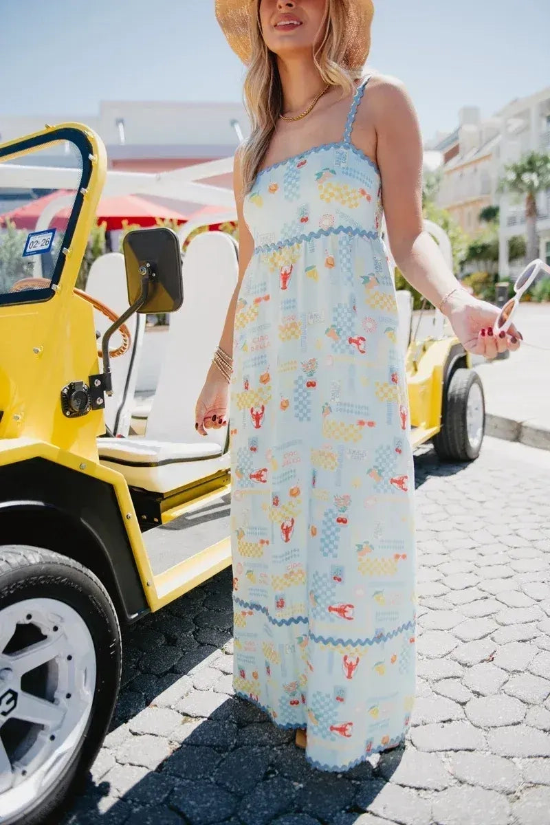 Woman in a straw hat standing next to a yellow jeep on a cobblestone street, wearing a light blue lobster and summer print maxi dress with blue ric-rac trim and spaghetti straps.