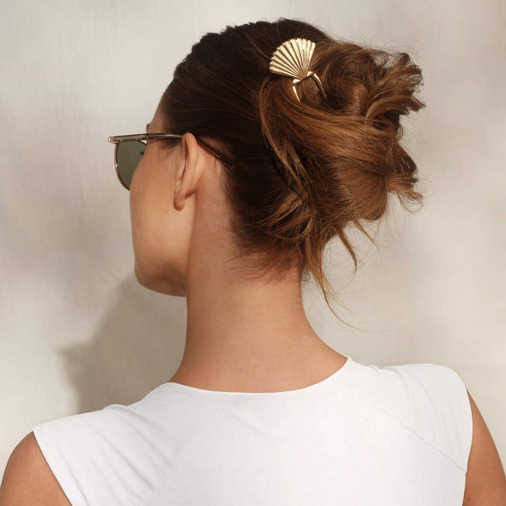 Back view of a woman with brown hair in a messy updo secured with a gold scallop shell hair pin, wearing sunglasses and a white top.