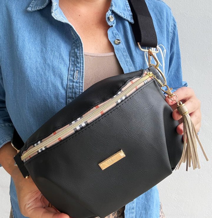 Woman holding black vegan leather belt bag with plaid-trim gold zipper, tassel charm, and wide black crossbody strap